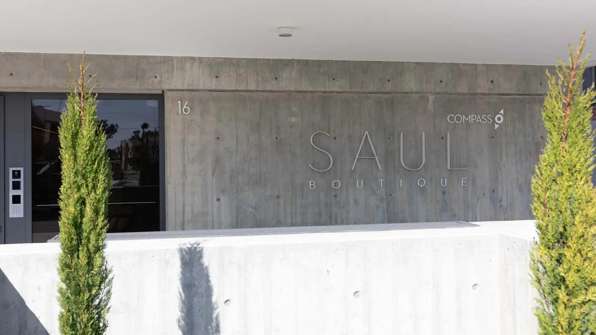 Entrance of the Saul Boutique residential building, with a concrete wall and cypress trees