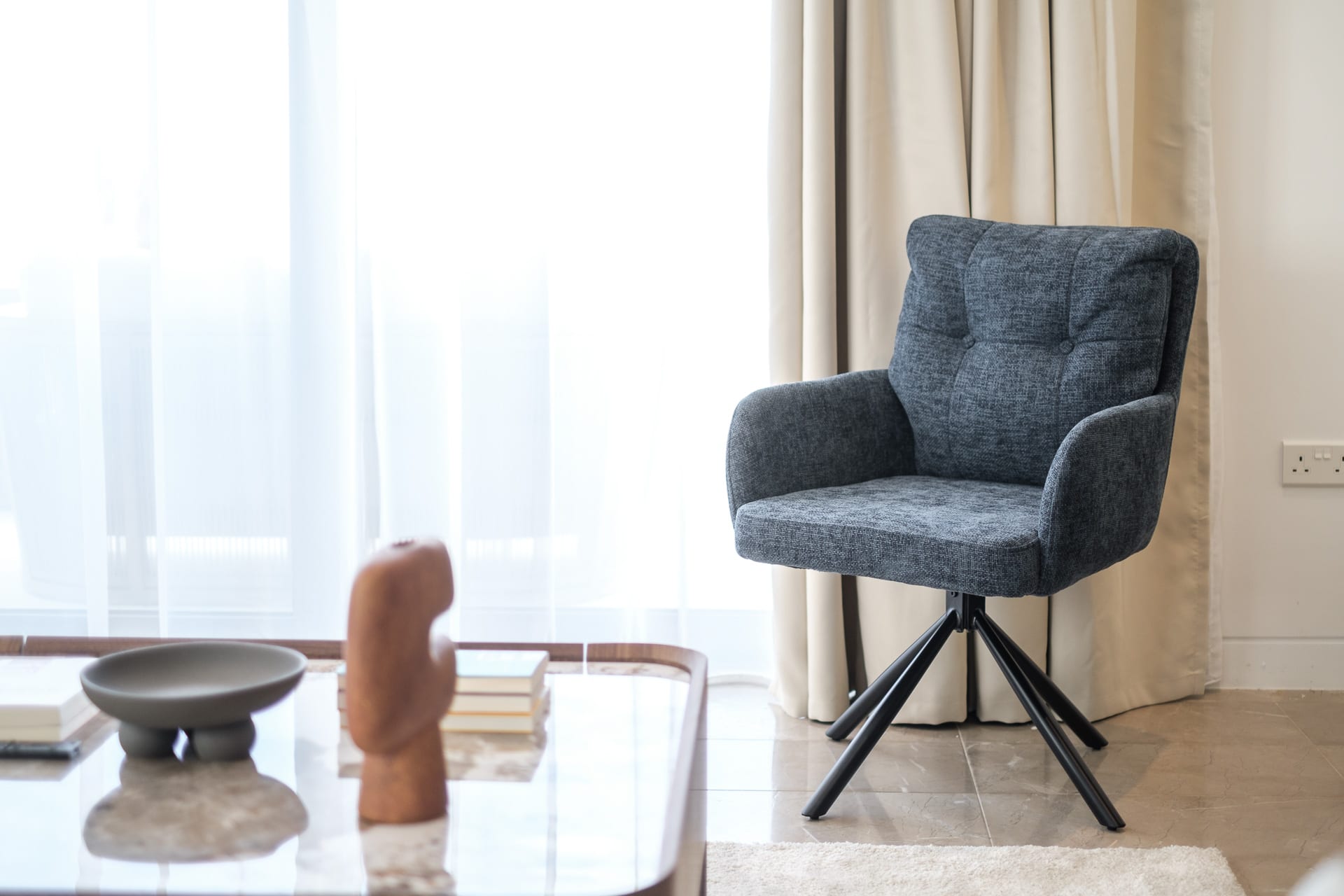Grey upholstered armchair by sheer curtains, with a sculpture and a bowl on a glass coffee table