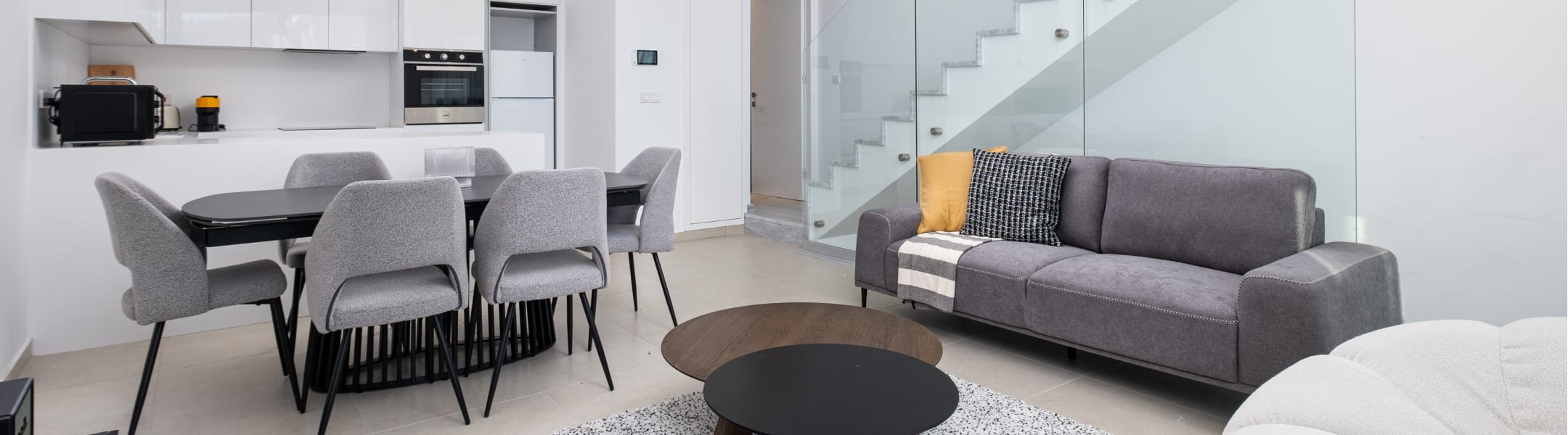 Open-plan apartment with a dining table, grey sofa, kitchen and glass-railed staircase