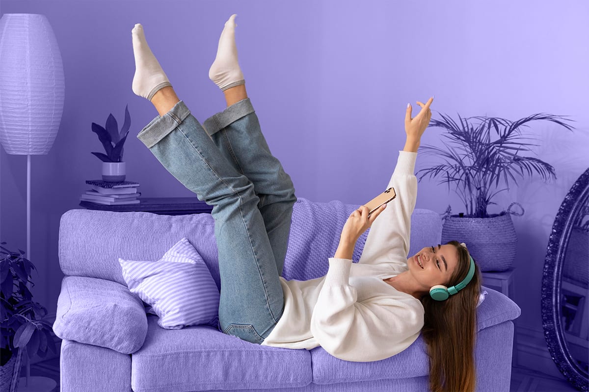 Woman lying on a sofa with her legs in the air, wearing headphones and holding a phone