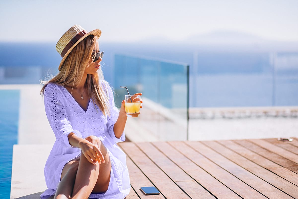 Woman in a straw hat holding a drink on a sunny poolside deck by the sea