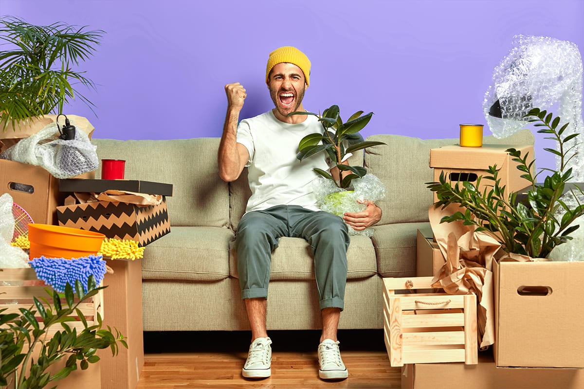 Excited man on a sofa holding a potted plant, surrounded by moving boxes